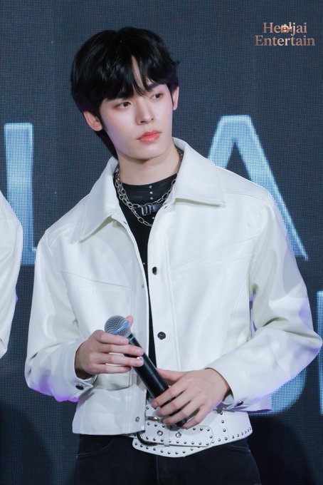 A young man with dark hair, wearing a white leather jacket with a studded belt and silver chain necklaces, holds a microphone while standing on stage. The "Heejai Entertain" watermark appears in the upper right corner, and the event branding in the caption corresponds to the "LABX GRAND OPENING DEXX THE EXPERIENCE" appearance.