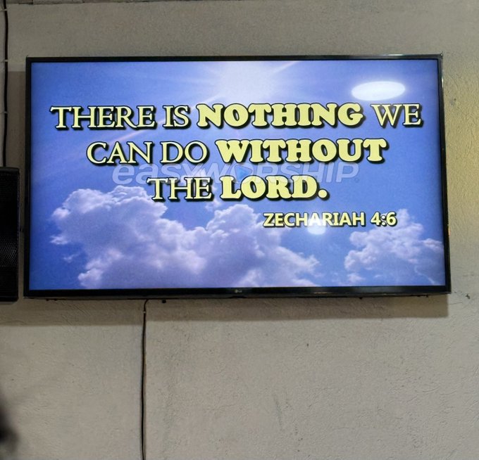A flat-screen TV mounted on a wall displays a Bible verse on a cloudy sky background, reading "THERE IS NOTHING WE CAN DO WITHOUT THE LORD. ZECHARIAH 4:6," with an "EasyWorship" watermark. The inspirational scripture aligns with the post's Sunday message encouraging faith, family time, and church attendance before starting the work week.