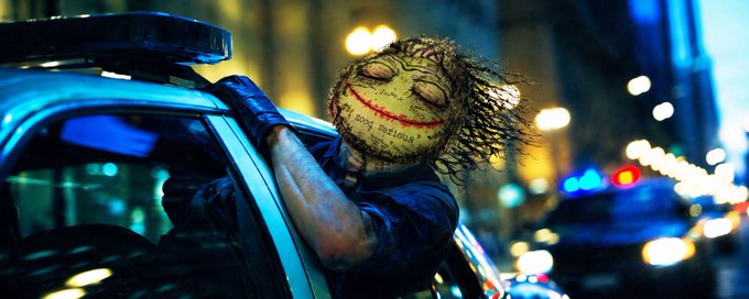 A cinematic shot of a masked figure wearing a creepy stitched doll-like mask with 'Why so serious?' written on it, leaning against a police car at night with blurred emergency lights in the background.