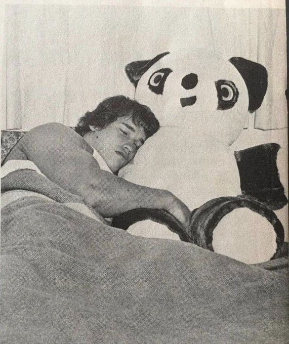 A black and white photo shows a shirtless man lying in bed next to a large stuffed panda bear.