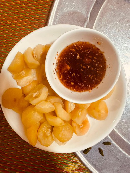 A photograph of a white plate containing sliced pickled fruits or vegetables arranged around a small bowl of dark dipping sauce with chili flakes, placed on a woven mat.