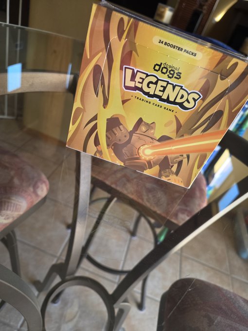 A box of "Dogmai Legends" trading card game containing 24 booster packs, sitting on a glass table with a character's image and flames on the cover.