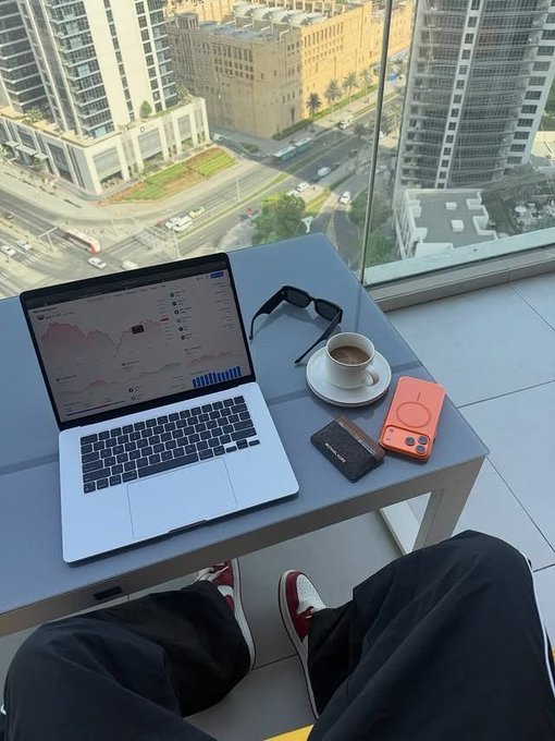 A person works on a MacBook displaying analytics dashboards and charts from a high-rise balcony overlooking a cityscape, with a coffee cup, sunglasses, an orange phone case, and a leather cardholder arranged on the table beside their red-and-white sneakers. The setup reflects the post's hustle mindset—grinding through gig work and analytics at the end of the month to chase earnings before the deadline hits.