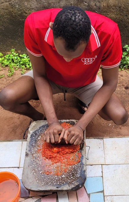 A young man in a red Audi-branded shirt squats on the ground, grinding red peppers on a stone slab outdoors.