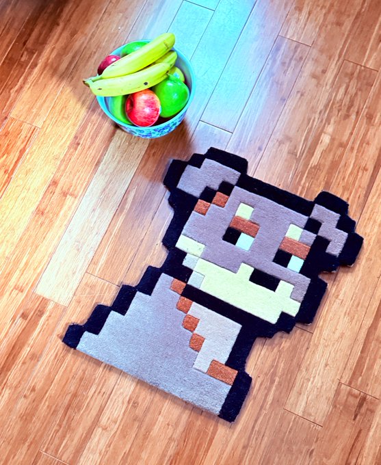 A pixel art rug of a gray and white bear with brown accents lies on a wooden floor next to a bowl of fruit containing bananas and apples.