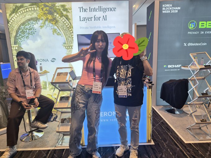 Two people posing at a blockchain convention booth labeled 'The Intelligence Layer for AI' and 'KOREA BLOCKCHAIN WEEK 2026' with '©BeldexCoin' and 'BCHAT' logos visible.