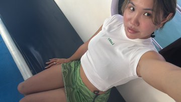 A selfie of a young woman wearing a white crop top with green text and a green watermelon-patterned skirt, posing indoors.