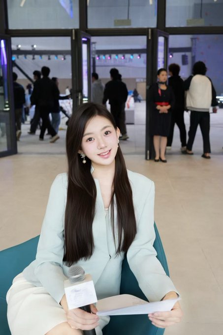 A young woman with long dark hair wearing a light blue blazer smiles at the camera while holding a microphone and papers at what appears to be an event venue.