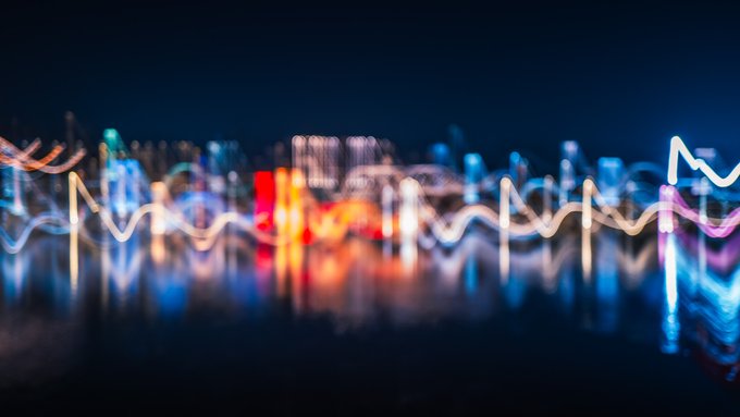 An abstract, blurred cityscape at night features vibrant light trails and bokeh effects in blue, red, white, and purple, creating a wave-like, neon aesthetic.