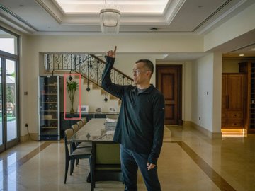 A man wearing glasses and a dark sweater stands in a spacious, well-lit home interior, pointing upward toward the ceiling near a dining area and staircase.