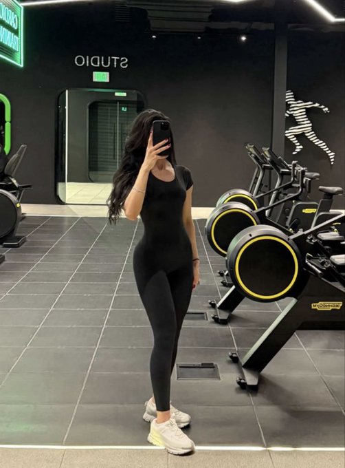 A mirror selfie taken in a gym (labeled 'STUDIO') shows a woman wearing a black short-sleeve jumpsuit and white sneakers, posing next to yellow Technogym stationary bikes.
