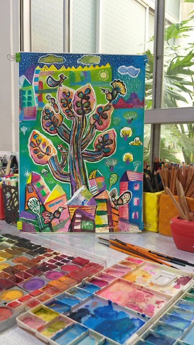 A vibrant, colorful painting of a stylized tree, birds, and houses sits on a table surrounded by watercolor palettes and paintbrushes near a window.