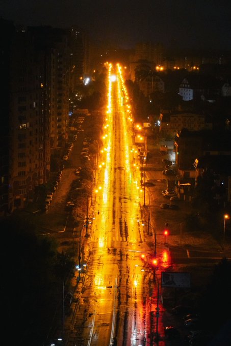 Aerial nighttime photograph of a long, illuminated city street lined with bright orange streetlights, with apartment buildings on either side.