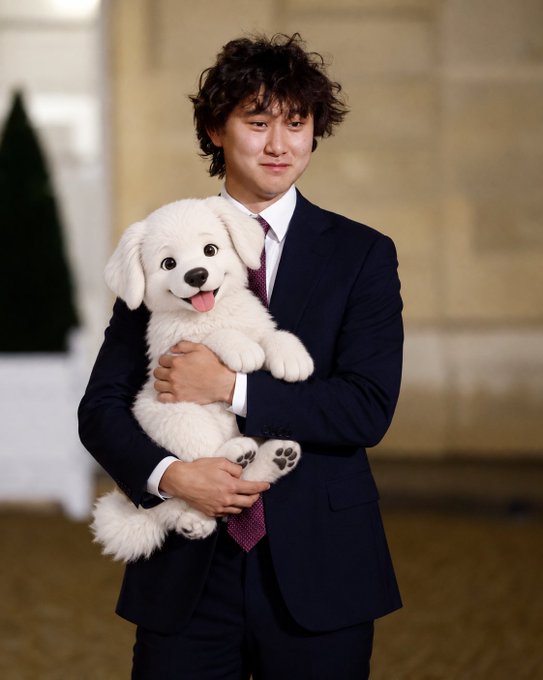 A young man with curly dark hair wearing a navy suit and purple tie holds a small white fluffy dog, with the dog's tongue sticking out.