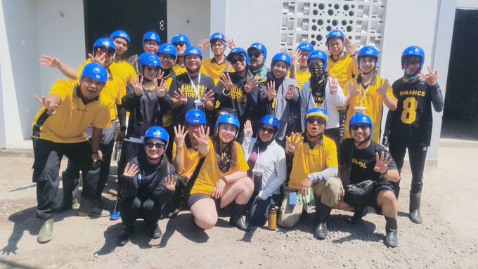 A large group of people wearing yellow shirts and blue helmets pose together outdoors, with one person sporting a black jersey reading "BINANCE 8," reflecting the post's celebratory shout-out to Binance Angels and SEA Angels as a top-notch community for building up together.