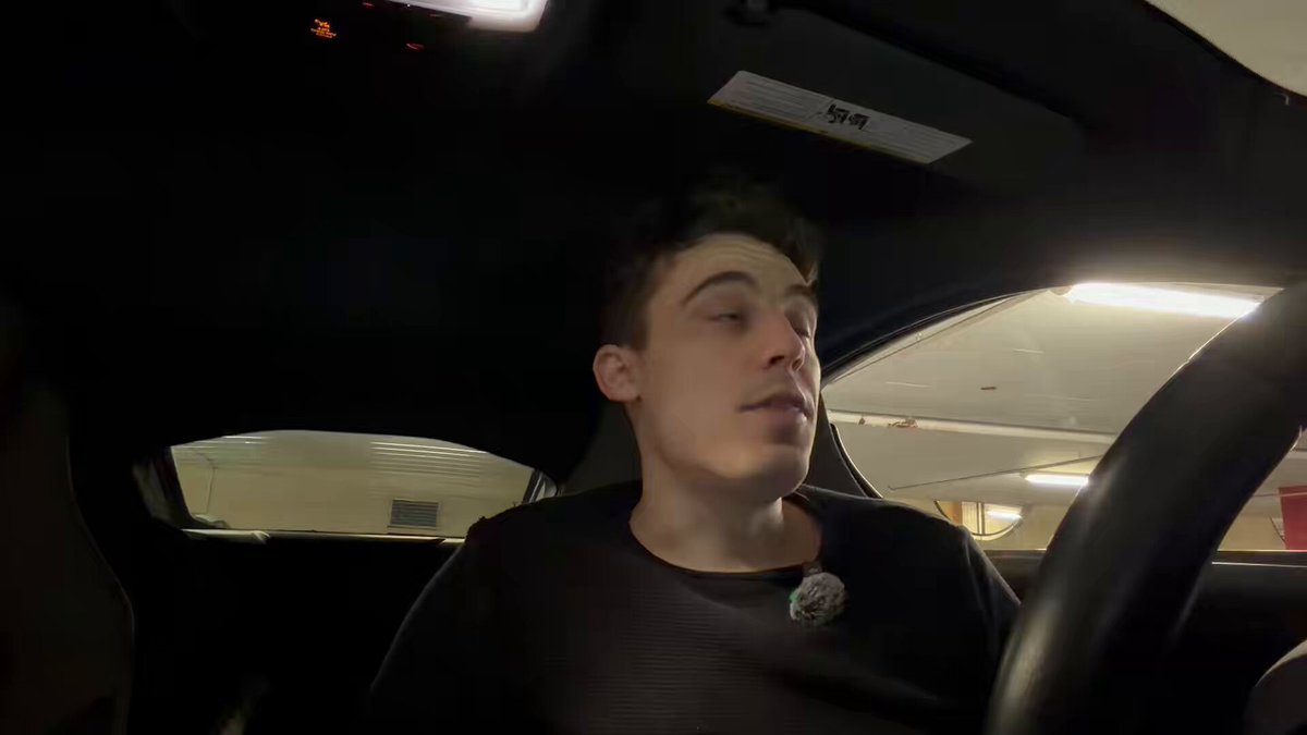 The image depicts a young man wearing a black shirt, sitting inside a car with his eyes closed and head resting on his hand while driving down the road at night. The scene appears to be captured from behind him as he drives through the night, creating an atmospheric mood.