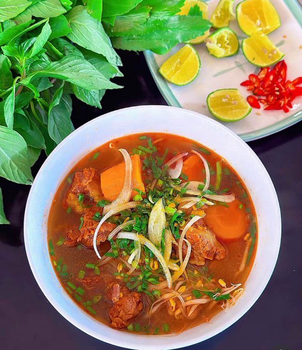 A bowl of Vietnamese-style beef stew (bò kho) garnished with onions, cilantro, and carrots, served alongside fresh basil, lime wedges, and sliced red chilies.