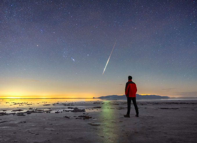 A person in a red jacket stands on a beach at night, watching a bright shooting star streak across a starry sky over the water.