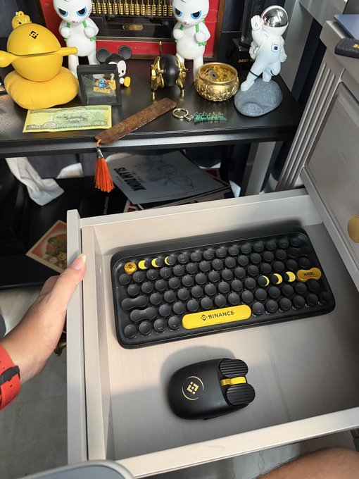 A person's hand opening a drawer to reveal a black and yellow Binance-branded wireless keyboard and a matching black mouse, with various figurines and memorabilia on the shelf above.