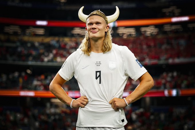 A digitally edited image of footballer Erling Haaland wearing an England #9 jersey and a Viking helmet with horns, smiling on the pitch at a stadium.