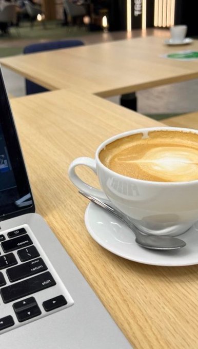 A close-up photograph of a cappuccino in a white cup on a saucer with a spoon, sitting on a wooden table next to a laptop keyboard in what appears to be a café setting.