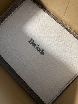 The image shows an open cardboard box containing a white item with black text that reads "DeGods". The box has been opened, revealing its contents inside. There is also a caption below the image that reads "Honestly if you still don't have a @DeGodsNFT I don't know what you are doing."