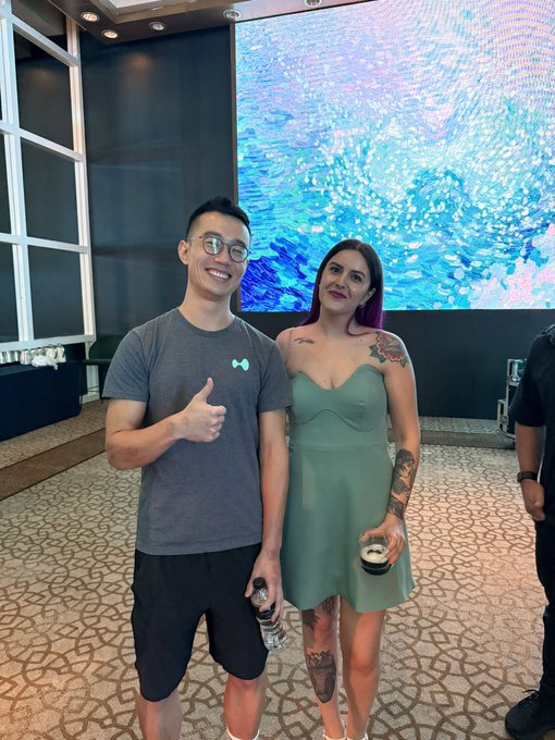 Two people pose together at an event, with the man on the left giving a thumbs-up while wearing a gray t-shirt and the woman on the right in a sage green dress with visible tattoos, holding a drink in front of a large blue-toned screen. A third person is partially visible on the right edge of the frame, suggesting a casual gathering related to the Hyperliquid project.