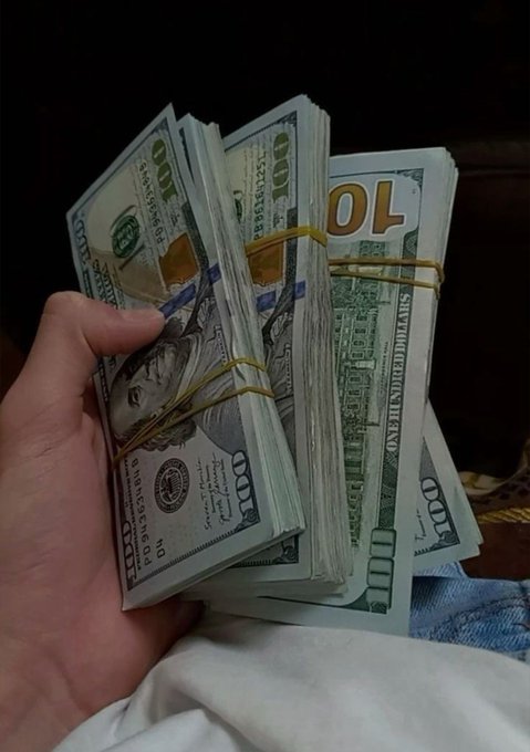 A hand holds several stacks of $100 bills bound by yellow rubber bands, with the caption celebrating another day of making money as a visual representation of earnings and hustle.