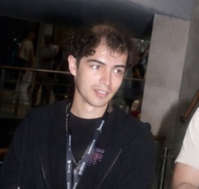 A candid photo of a young man with curly dark hair wearing a black jacket and graphic t-shirt, posing at what appears to be an event.