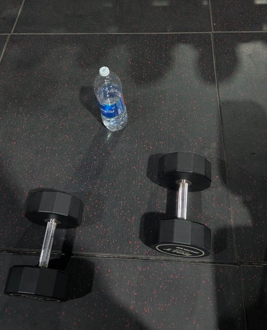 Two black dumbbells and a partially consumed water bottle rest on a speckled black gym floor, capturing the moment just after a workout. The water bottle's blue "Nestlé Pure Life" label is visible, symbolizing the post-workout hydration break before the day begins.
