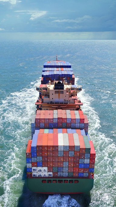 An aerial view from the stern of a large cargo ship stacked with colorful shipping containers sailing through blue ocean waters under a partly cloudy sky.