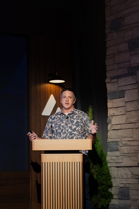 A man in a blue and white floral shirt stands at a wooden podium with a stylized 'M' logo on the wall behind him, gesturing while speaking at an indoor event.