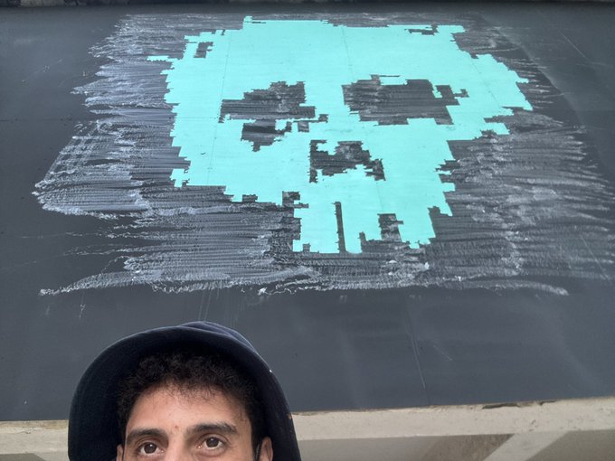 The image features a man wearing a black hat, standing next to a large piece of artwork that depicts a skull made out of tiles or blocks. The artwork appears to be in progress as there are some areas where the artist has added more details or colors. The background is dark and somewhat blurry, which makes it difficult to see other objects around the man. The image also contains text that reads "work in progress Look at it closely featuring @XCOPYART powered by @opensea @NFCsummit".