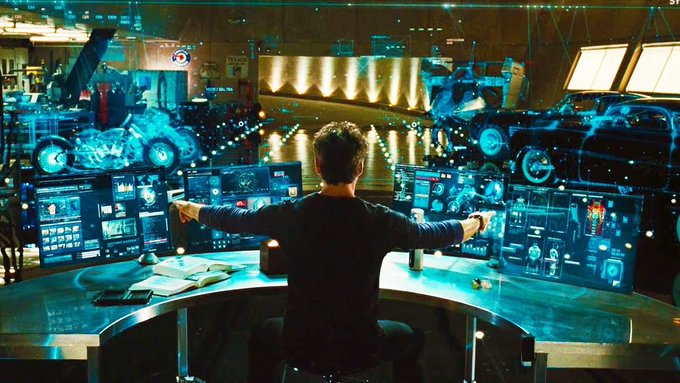 A man viewed from behind sits at a curved desk with multiple monitors, surrounded by holographic blue displays in a futuristic, high-tech control room.