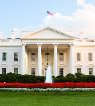 The image shows the North Portico of the White House in Washington, D.C., featuring its iconic columns, the American flag flying above, and a fountain surrounded by red flowers in the front garden. This iconic backdrop directly ties to the post, as it sets the scene for the upcoming meeting between White House officials and Bitcoin and crypto market executives, signaling a high-level policy discussion on the future of digital assets in the United States.