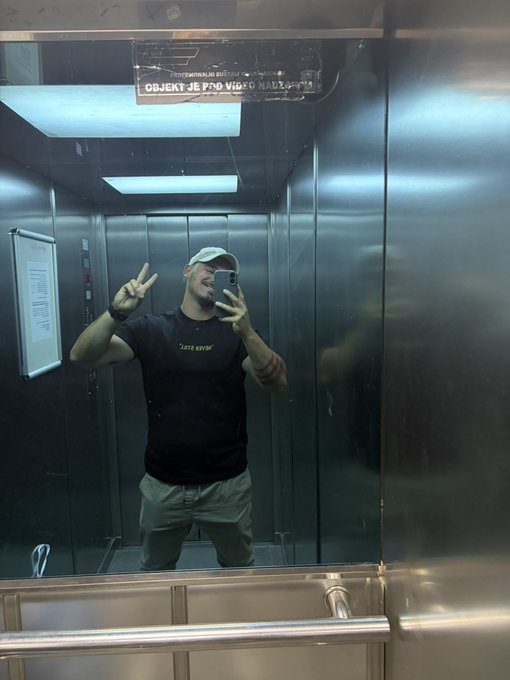 A mirror selfie of a bearded man wearing a cap and black t-shirt making a peace sign inside a metal elevator, with a sign reading 'OBJEKT JE POD VIDEO NADZOROM' visible above.