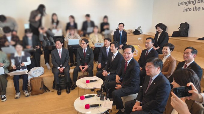A group of formally dressed South Korean politicians, likely members of the People Power Party, are seated together at a press event or meeting, with microphones placed on small tables in front of them. The setting appears to be an institutional or governmental building, and the gathering likely relates to legislative discussions, possibly including the proposed postponement of virtual asset taxation mentioned in the post.