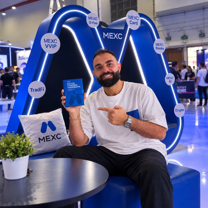 A smiling bearded man sits on a blue MEXC-branded bench at TOKEN2049, holding up the "MEXC Stock Trading Handbook" and pointing at it, surrounded by circular badges promoting MEXC's services (MEXC Card, Stocks, AI, Crypto, TradFi, VVIP, 0-Fee). The image promotes the post's offer of a free handbook to qualifying traders at the event, with the MEXC branding and hashtags (#MEXCTradFi, #MEXC0FEE) reinforcing the campaign.