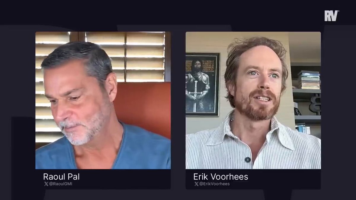 The image shows two men sitting next to each other, with one man wearing a blue shirt and the other wearing a white shirt. They are both looking at something off-camera, possibly engaged in a conversation or watching a video together. The background of the image is dark, which makes it difficult to see any text or additional objects.