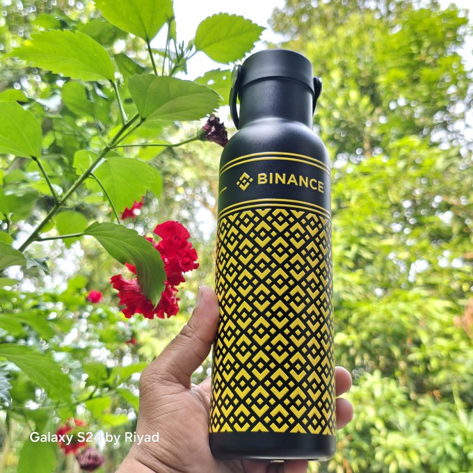 The image features a hand holding up a black and yellow water bottle with the word "Binance" written on it, surrounded by red flowers. The background of the photo includes trees, suggesting that this scene takes place outdoors or in a natural setting. The caption provided connects the image to a post about Web3 and crypto, which might be related to the water bottle's brand or features.