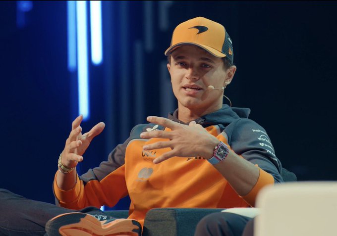A man wearing an orange McLaren racing cap and orange jacket gestures with his hands while speaking at what appears to be a conference, wearing a headset microphone.
