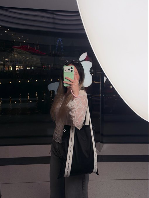 A young woman with long blonde-highlighted hair takes a mirror selfie in front of an Apple Store window, holding a mint green iPhone while wearing a white lace top and carrying a black tote bag.
