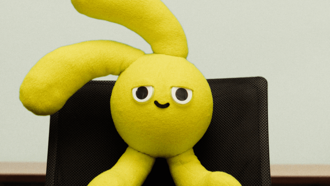 A yellow plush toy with long ears and a smiling face sitting on a black mesh chair.
