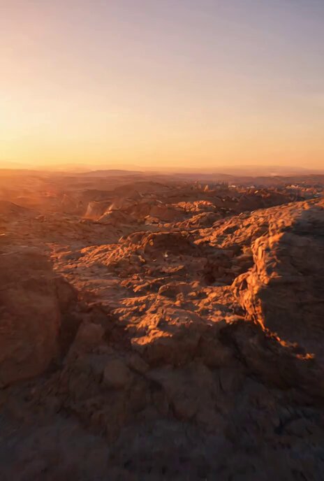 The image shows a breathtaking view of a desert landscape at sunset. The sky is painted with warm hues of orange and yellow, creating a stunning backdrop for the rugged terrain below. The sun's rays are shining down on the rocky mountainside, casting long shadows and highlighting the textures of the rocks. The perspective of the photo is taken from a high angle, providing an expansive view of the desert landscape that stretches out into the distance.