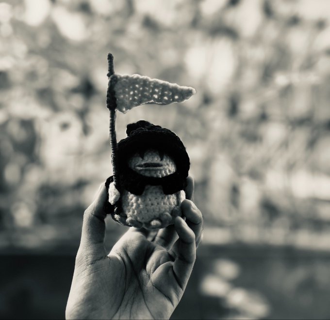 A black and white photograph of a hand holding a small crocheted figure holding a flag against a blurred natural background.