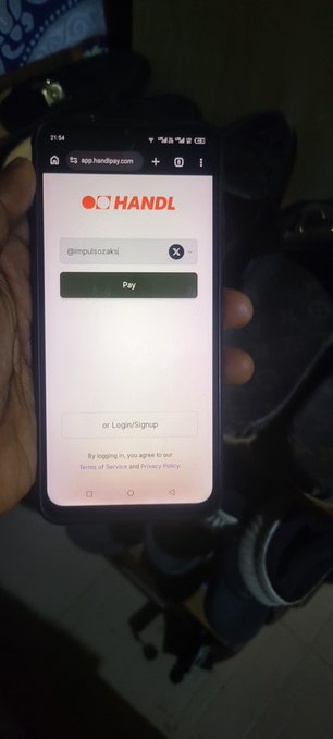 A hand holds a smartphone displaying the HANDL app login screen with the username '@mpufosozaki' filled in, showing buttons for 'Pay' and 'or Login/Signup' on a dark background with shoes visible below.