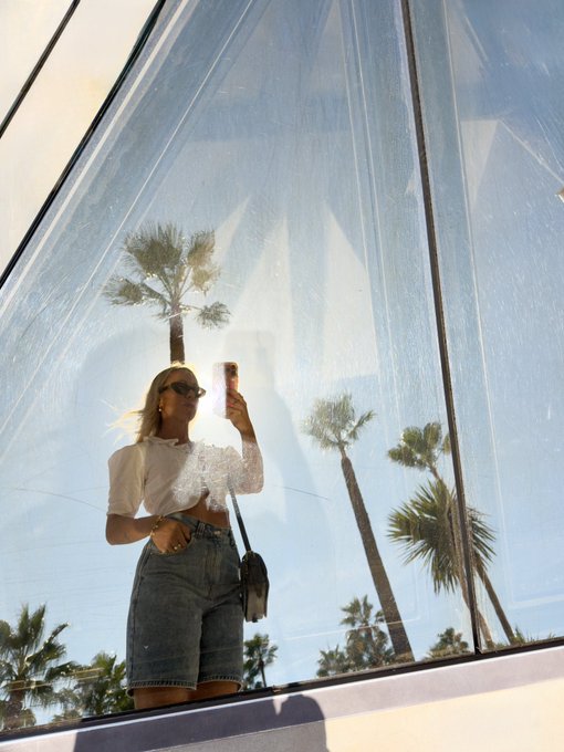 A blonde woman wearing sunglasses, a white crop top, and a denim skirt takes a mirror selfie with her phone, with palm trees and bright sky reflected in the glass behind her.
