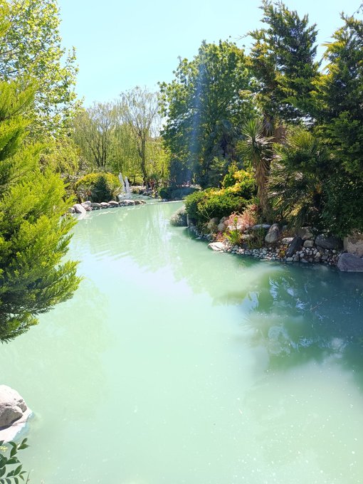 A scenic view of a turquoise pond surrounded by lush green trees, shrubs, and rocks in a landscaped garden under a bright blue sky.