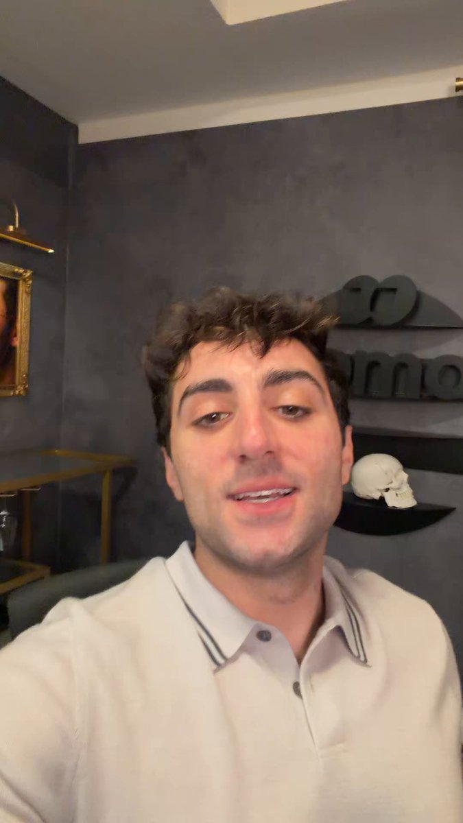 The image features a man wearing a white polo shirt, smiling at the camera while taking a selfie with his phone. There are two other people visible in the background, but they are not the main focus of the photo. The caption "Saylor was just testing the sell button..." suggests that someone has posted this photo on social media to share their experience or to promote something related to the image's content.

The man is wearing a white polo shirt with black stripes and appears to be in a room with gray walls, which could indicate an indoor setting like a living room or a bedroom. The presence of two other people in the background might suggest that they are also taking photos or participating in some sort of event related to the image's content.