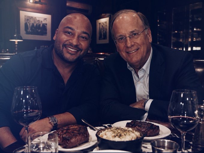 Two men in dark blazers smile together at an upscale steakhouse, with plates of steak, a side dish, and wine glasses set before them. The post references their meeting at Davos, contrasting the candid-looking photo with skepticism that it could be AI-generated.